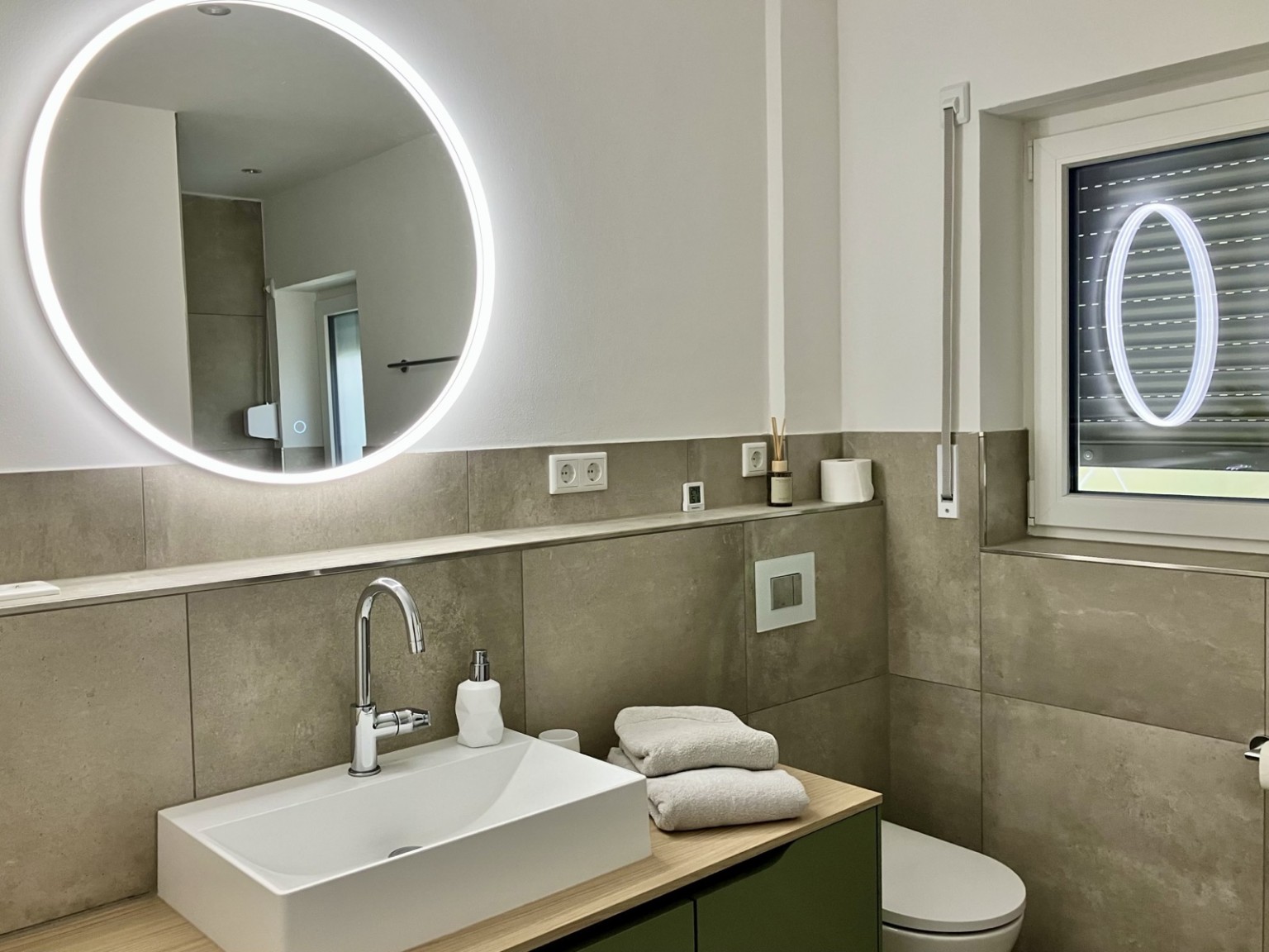 Zum Vergrößern anklicken Modernes Badezimmer mit Waschtisch, beleuchtetem Rundspiegel, WC und Fenster